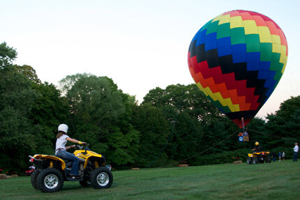 Person on yellow ATV driving towards rainbow-patterned hot air balloon in a field